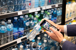 A hand holding a mobile phone, using the phone to scan a barcode on a bottle of water to make payment.