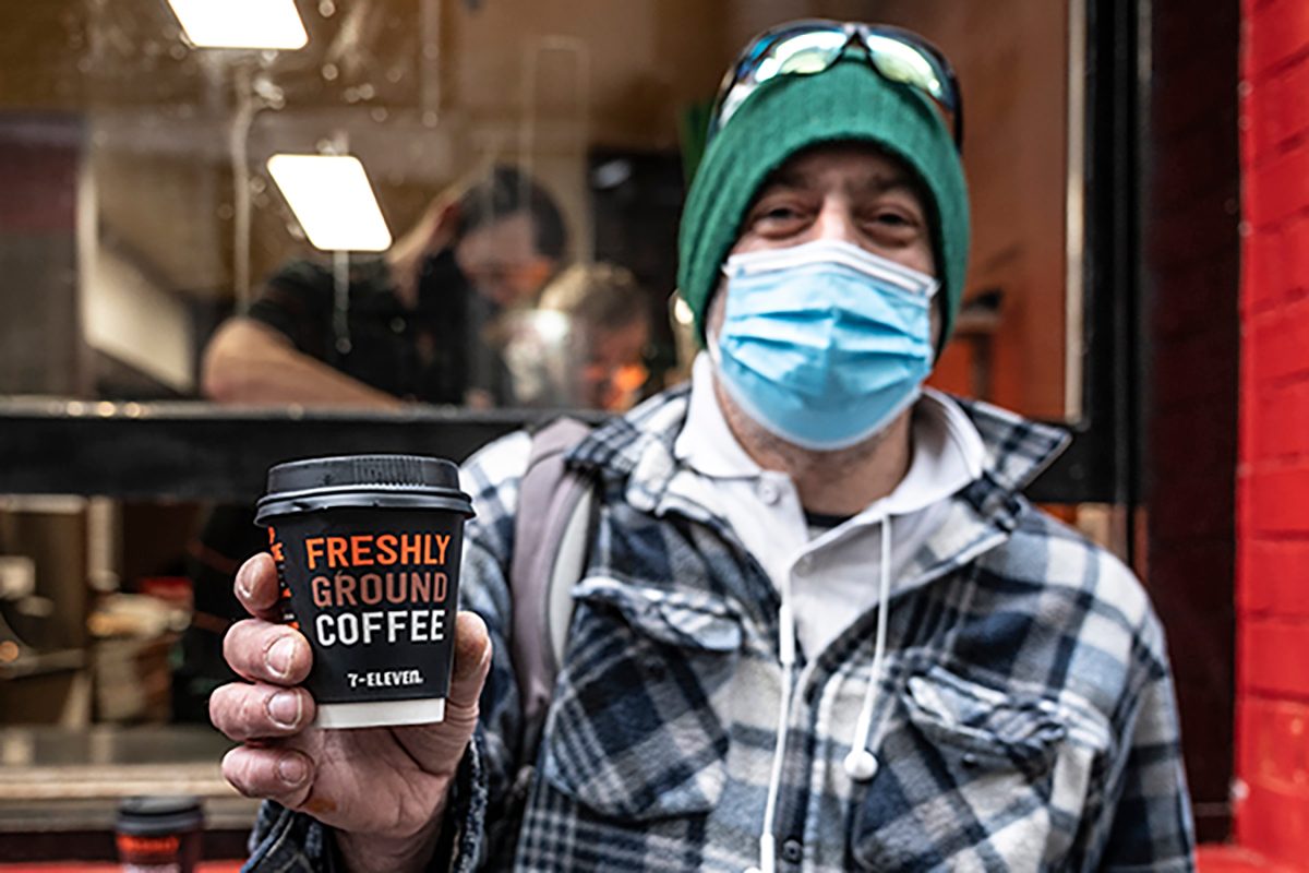 A person wearing a flanalette shirt, a beanie and a mask. Holding a cup of 7-Eleven coffee and a sandwich.