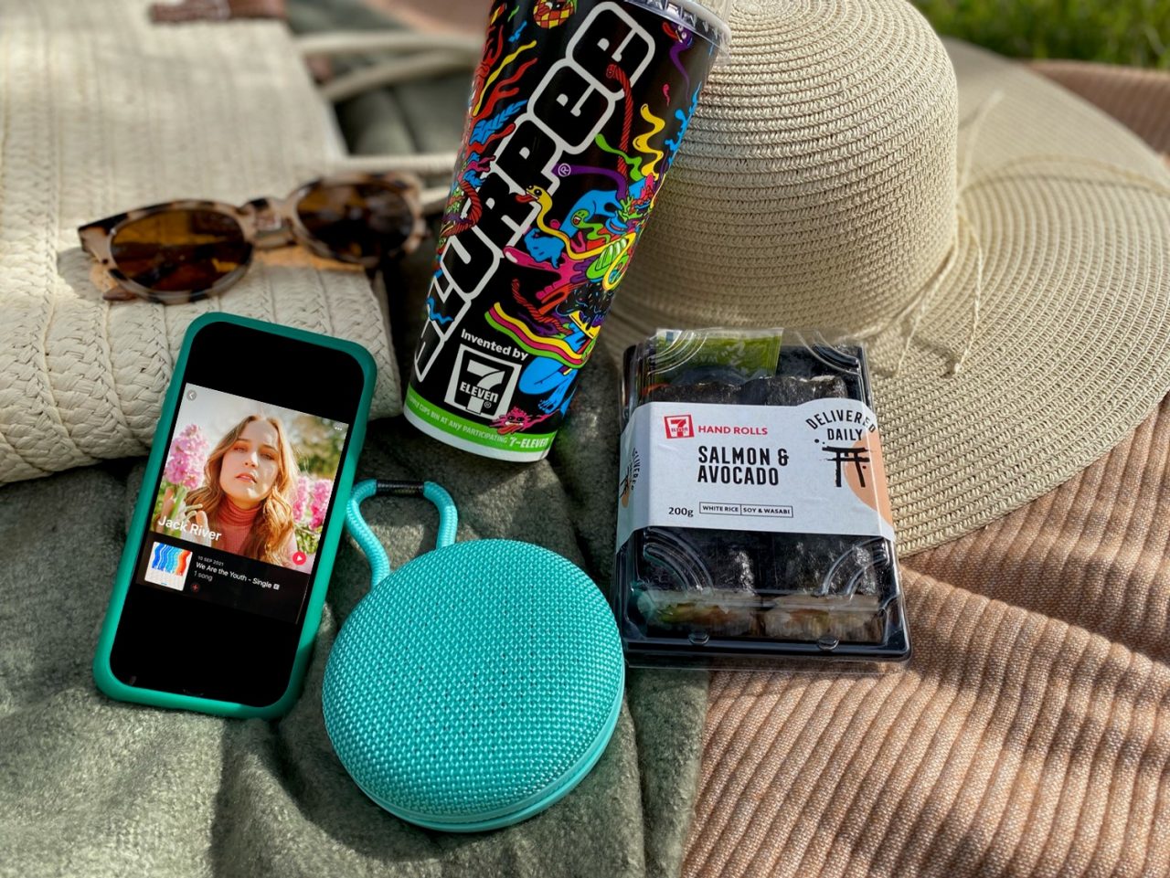 A picture of a picnic blanket with a summer hat, sunglasses, a Slurpee Cup, a pack of 7-Eleven sushi, a portable music speaker and an iphone playing music. Artist Jack River's single is pictured in the screen.