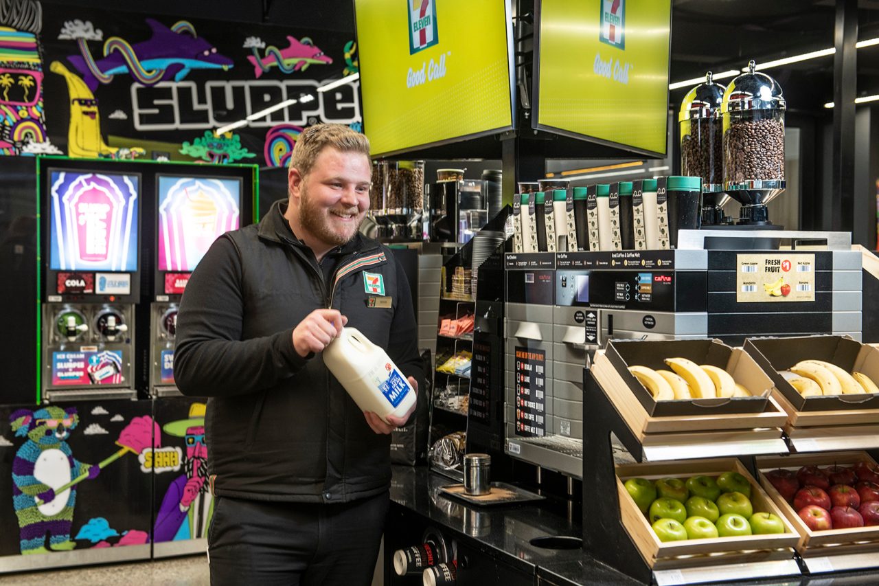 A picture of a man smiling in a 7-Eleven uniform, filling up milk in a coffee machine in a 7-Eleven store.