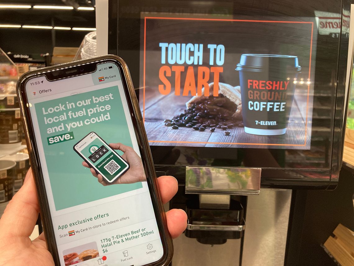 Picture of a phone with My 7-Eleven App fuel price lock on the screen in front of a 7-Eleven Coffee Machine.