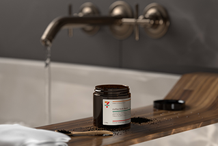 Picture of a tub of 7-Eleven coffee scrub, sitting on a wooden bath shelf, tap running in background.
