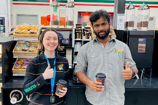 Emily and Ram from 7-Eleven Delacombe in their uniforms, smiling, in front of coffee machine.