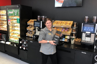 Casey Reece is standing in front of a 7-Eleven Coffee machine, in her store uniform and holding a bag of coffee beans.