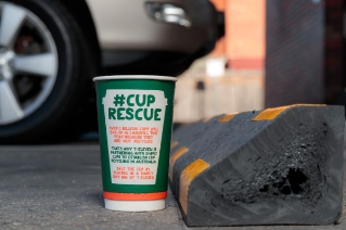 A 7-Eleven coffee cup with #CupRescue information on it, sitting on the ground next to a car park bumper made from recycled coffee cups.