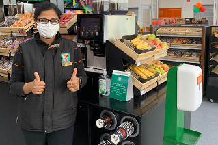 A female team member wearing a 7-Eleven uniform. She is wearing a mask, standing next to a hand sanitiser unit. She has her thumbs up. 